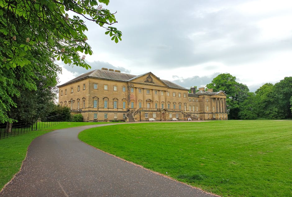 A large historic brick building with multiple windows and white decorative accents, situated in a landscaped area with a gravel pathway leading up to the entrance. The facade features symmetrical desi