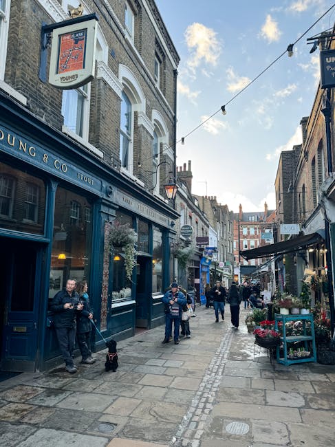 A narrow residential street in Hampstead with cobblestone paving and a slight curve, lined with traditional brick buildings featuring white window frames and black iron railings. There are small potte